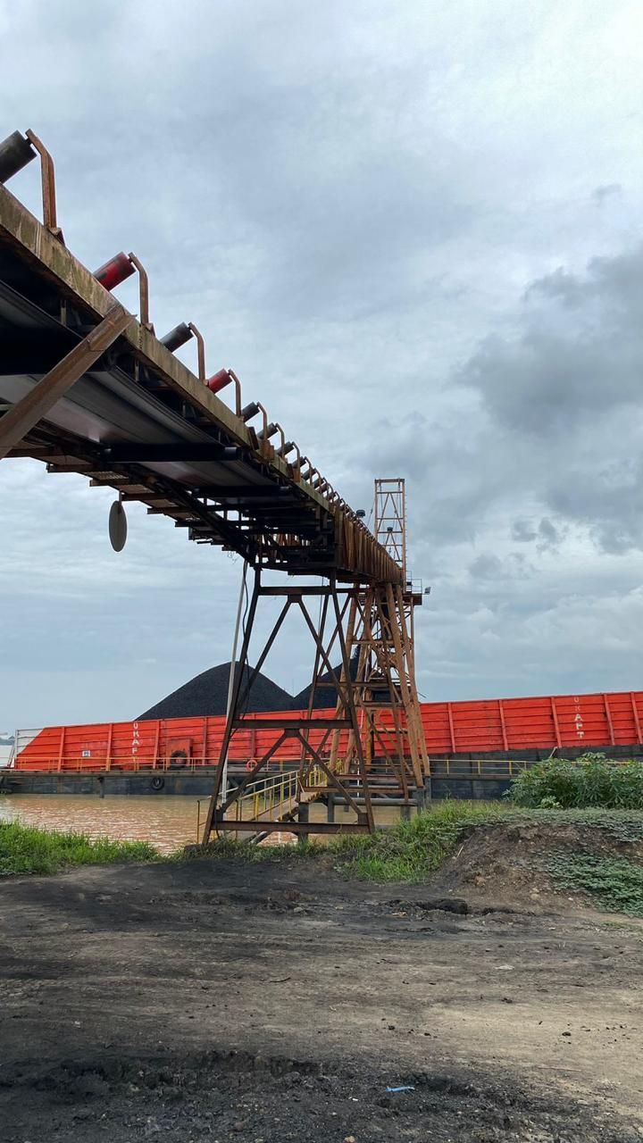 Kegiatan penumpukan dan pengiriman batubara via conveyor di tepi sungai.